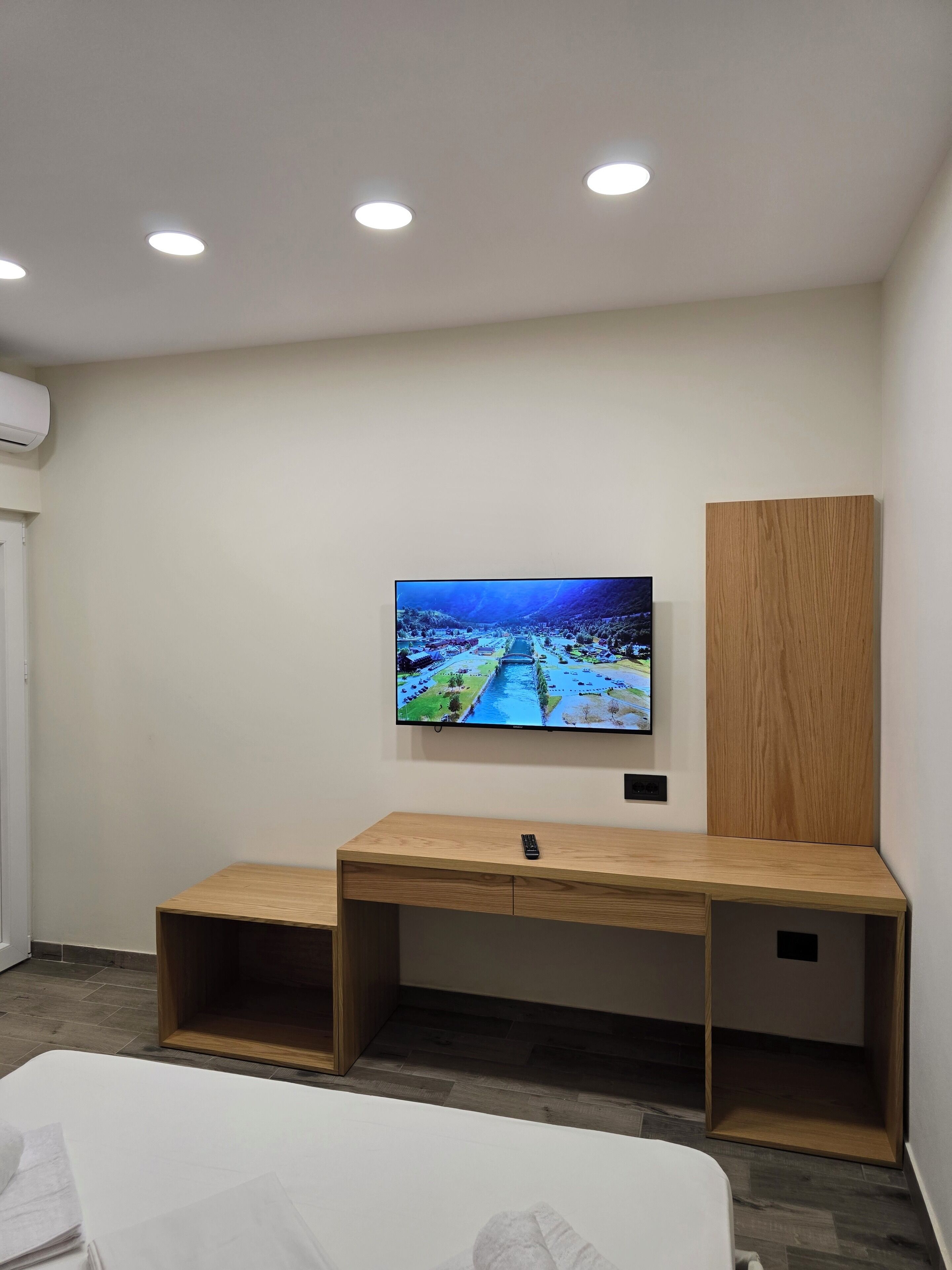 A modern room with a flat-screen TV mounted on the wall, a wooden desk, and a bed with a white mattress.
