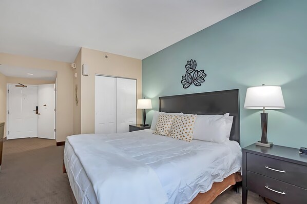 A hotel room with a large bed, a nightstand with a lamp, a wall-mounted artwork, and a door leading to another room.