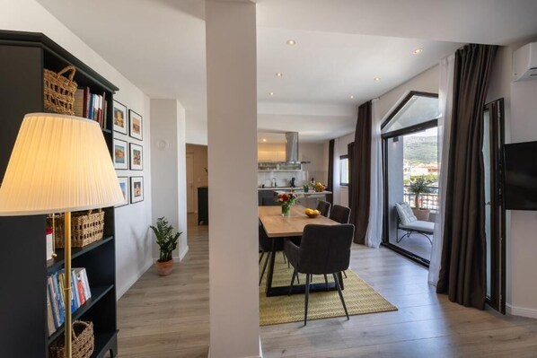 A modern living room with a dining area, a kitchen in the background, and a large window with curtains.