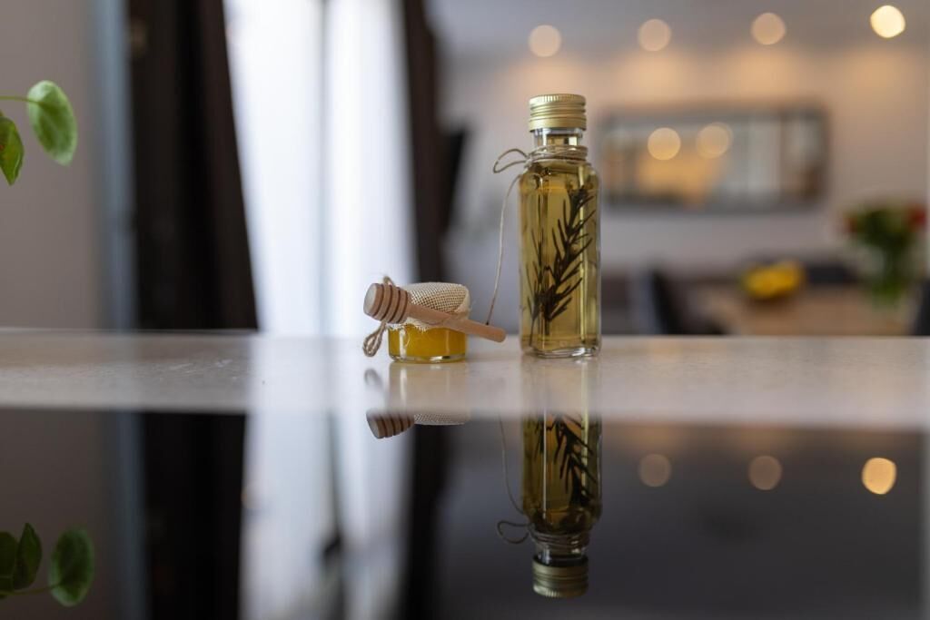 Une bouteille contenant un liquide, accompagnée d’une cuillère à miel en bois et d’un petit pot de miel, le tout posé sur une surface réfléchissante.