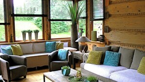 Ein gemütliches Wohnzimmer mit einer Couch, Sesseln, einem Couchtisch und Blick auf Grün hinter großen Fenstern.