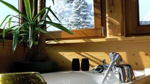 Innenansicht einer Holzhütte mit Badewanne, einer Topfpflanze und Blick auf verschneite Bäume.