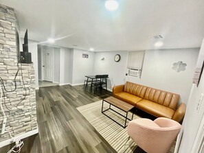 Un salon moderne avec un canapé en cuir marron, un fauteuil rose, une table basse en bois et une table à manger avec des chaises. On y trouve également un mur d’accent en pierre, une horloge et un climatiseur fixé au mur.