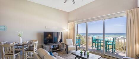 A spacious living room with a large TV, comfortable seating, a dining area, and a balcony with a view of the beach.