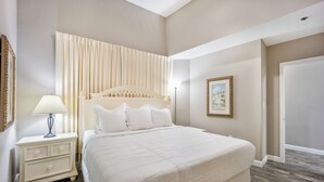 Een slaapkamer met een groot bed, een nachtkastje met een lamp, een lijst met een foto aan de muur en een deur aan de rechterkant.