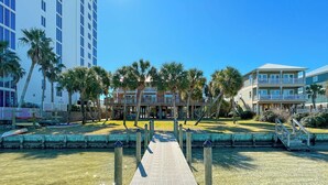 A waterfront hotel with a dock leading to a building with multiple balconies, surrounded by palm trees and clear blue skies. - Steps to the beach and Dock on the Bay! Pool, family and pet friendly! (Gulf Shores)
