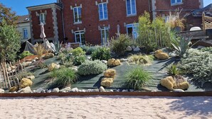 Ein angelegter Garten mit verschiedenen Pflanzen und Steinen, einem Holzzaun und einem Gebäude mit zahlreichen Fenstern im Hintergrund.