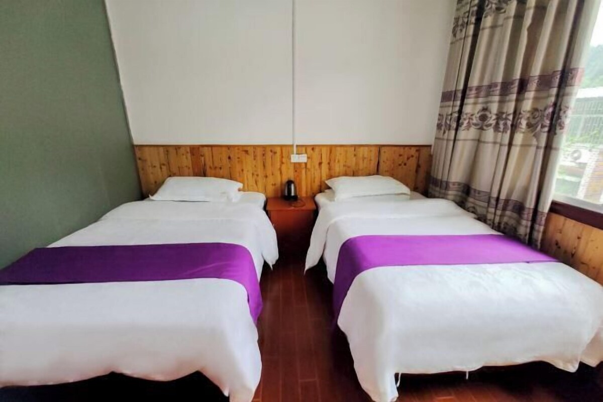 Une chambre avec deux lits, chacun recouvert d’une courtepointe blanche et violette, doté d’une tête de lit en bois et agrémenté d’une table de chevet avec une bouilloire.
