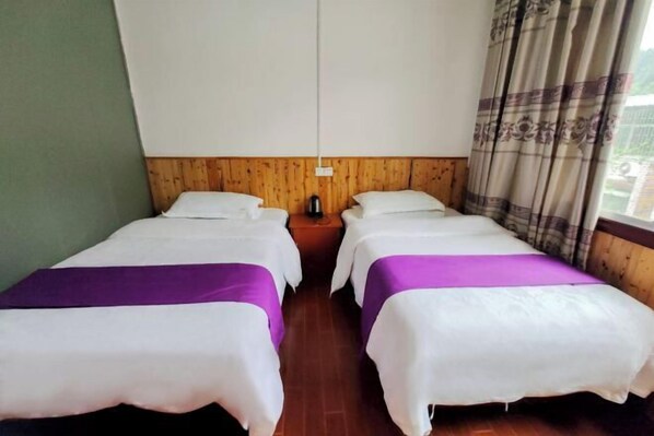 Une chambre avec deux lits, chacun recouvert d’une courtepointe blanche et violette, doté d’une tête de lit en bois et agrémenté d’une table de chevet avec une bouilloire.