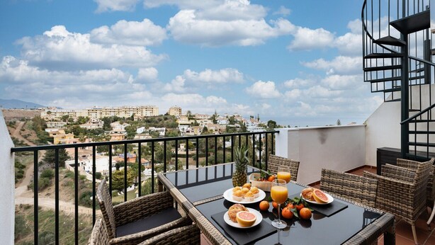 A balcony with wicker furniture, a table set with breakfast, and a view of a town. - Faro's Angel Miraflores Penthouse (Mijas)