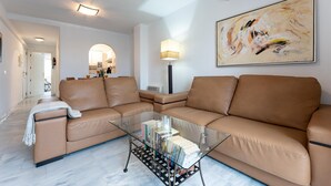 A modern living room with a brown leather sofa, a glass coffee table, and a large abstract painting on the wall. - Faro's Angel Miraflores Penthouse (Mijas)