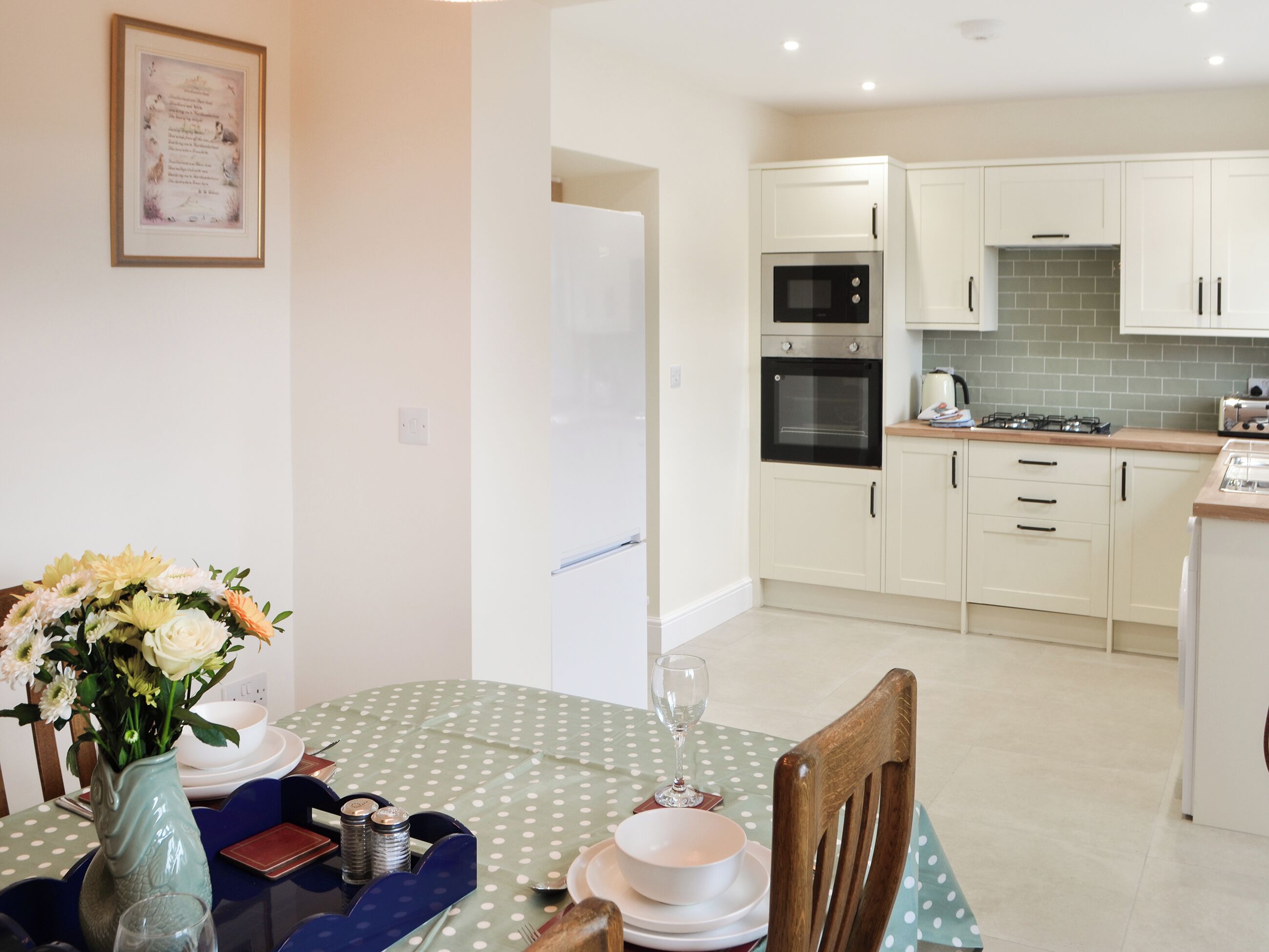 A modern kitchen with white cabinets, a built-in oven, and a microwave. There is a dining table with a polka dot tablecloth and a vase of flowers.