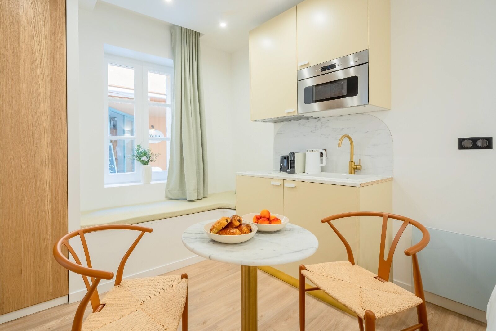 Een compacte keuken met een ingebouwde oven, een spoelbak en een raam met gordijnen. Er staan twee houten stoelen bij een kleine ronde tafel met een schaal fruit.