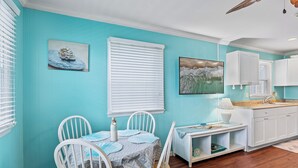 A dining area with a table set for two, white chairs, a white cabinet, and a kitchen area with white cabinets and a countertop. - Harbor Breeze 53 by Seaside Vacations & Sales (Ocean City)