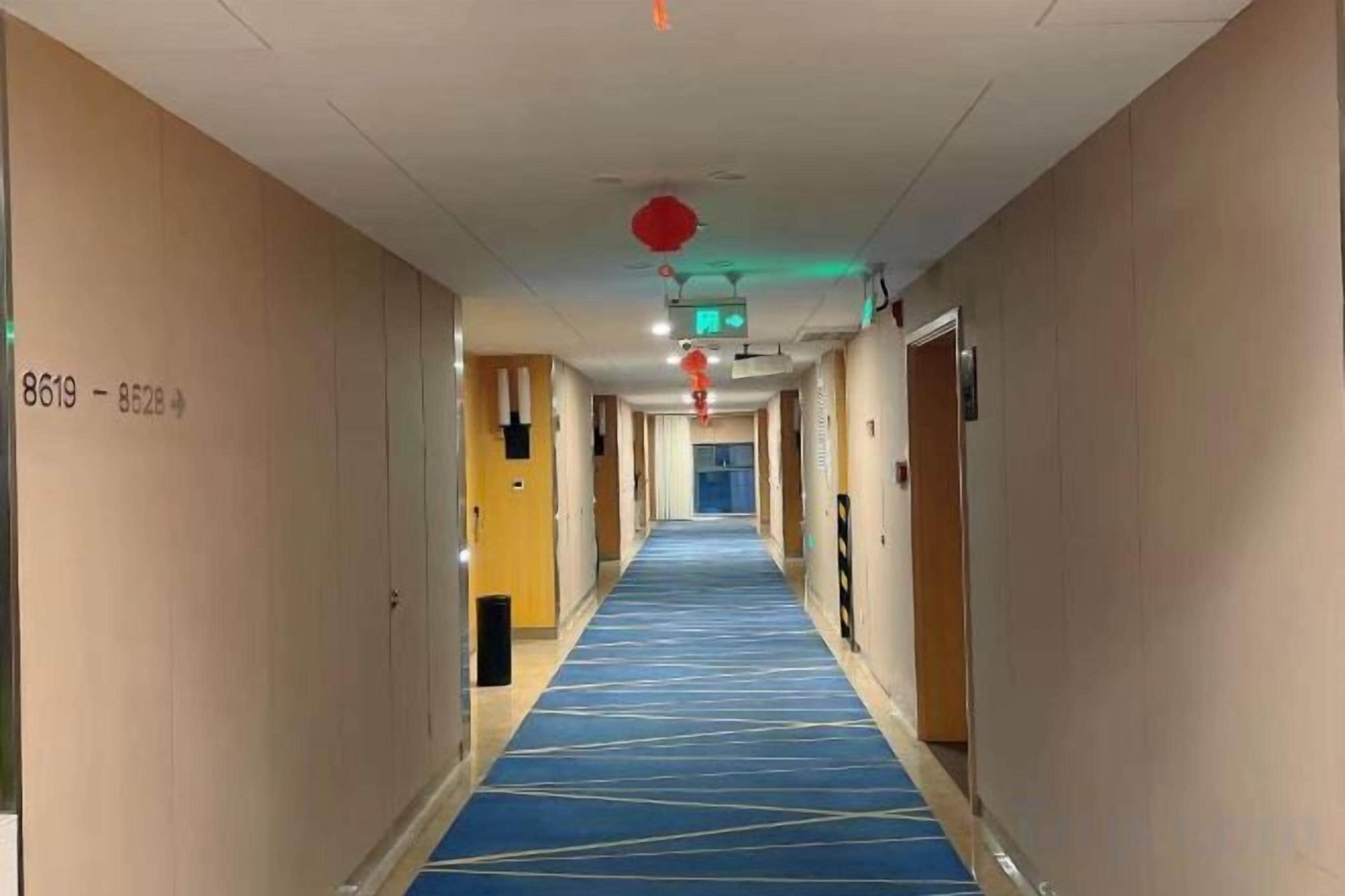 Pasillo de hotel con alfombra azul, puertas a ambos lados y un letrero de salida rojo en el techo.