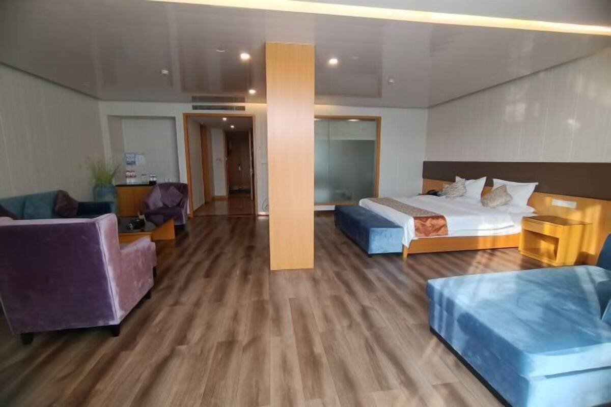 Ein modernes Hotelzimmer mit Holzboden, einem Bett mit blauer Kopfteil, einer Sitzecke mit violettem Sofa und einem Nachttisch aus Holz.