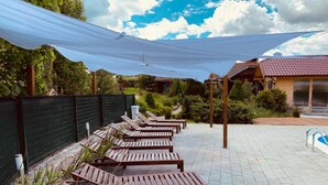 A row of wooden lounge chairs under a shaded area with a tiled patio, surrounded by greenery and a building with a red-tiled roof. - Villa Rubcic (Rakovica)