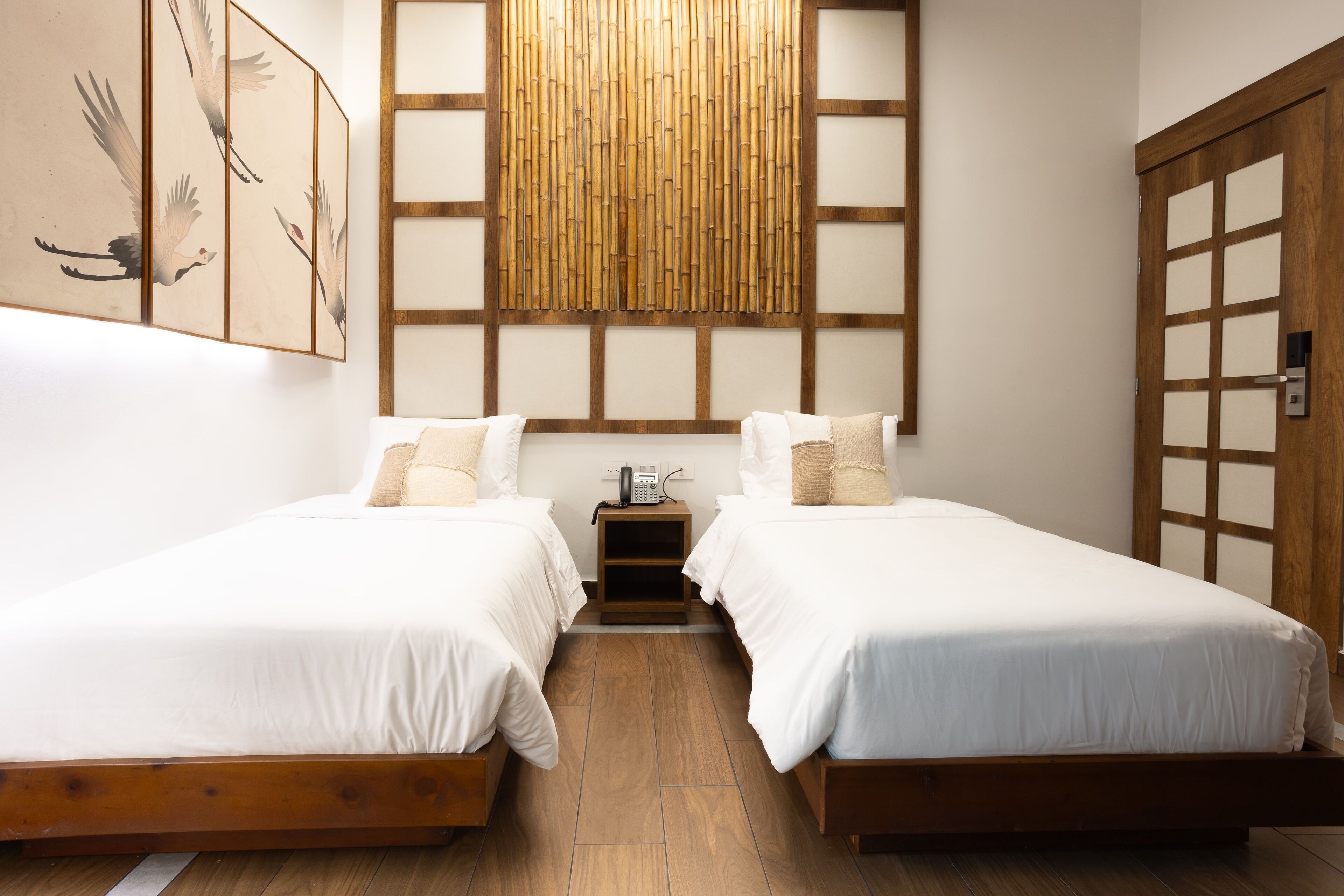 A room with two single beds, a wooden floor, a wall-mounted telephone, and a wooden door with glass panels.