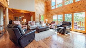 Ein großzügiges Wohnzimmer mit Kamin, Ledersesseln, einer Sofagarnitur und großen Fenstern mit Blick ins Freie.