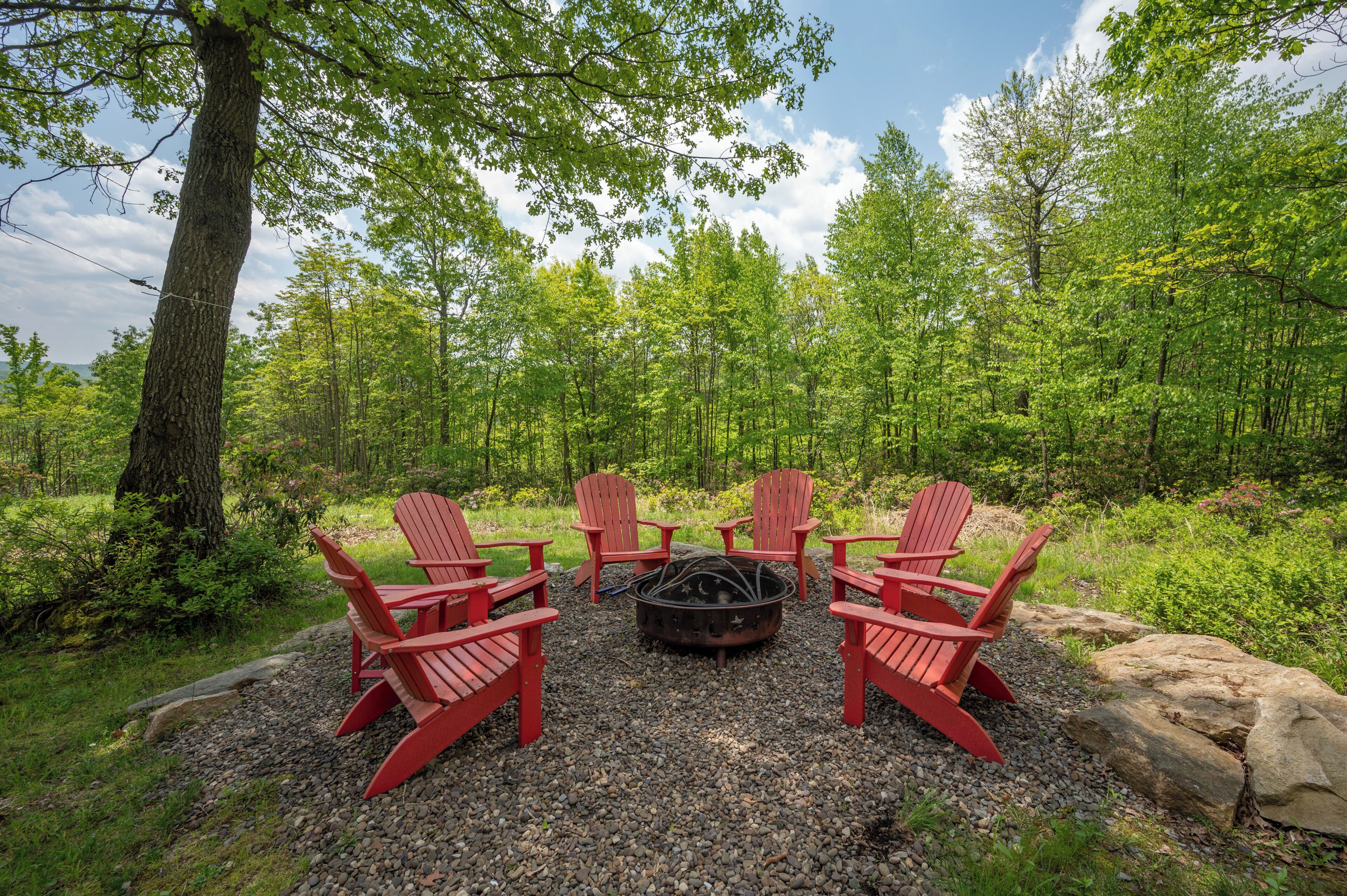 A forest setting with a fire pit and red Adirondack chairs arranged around it.