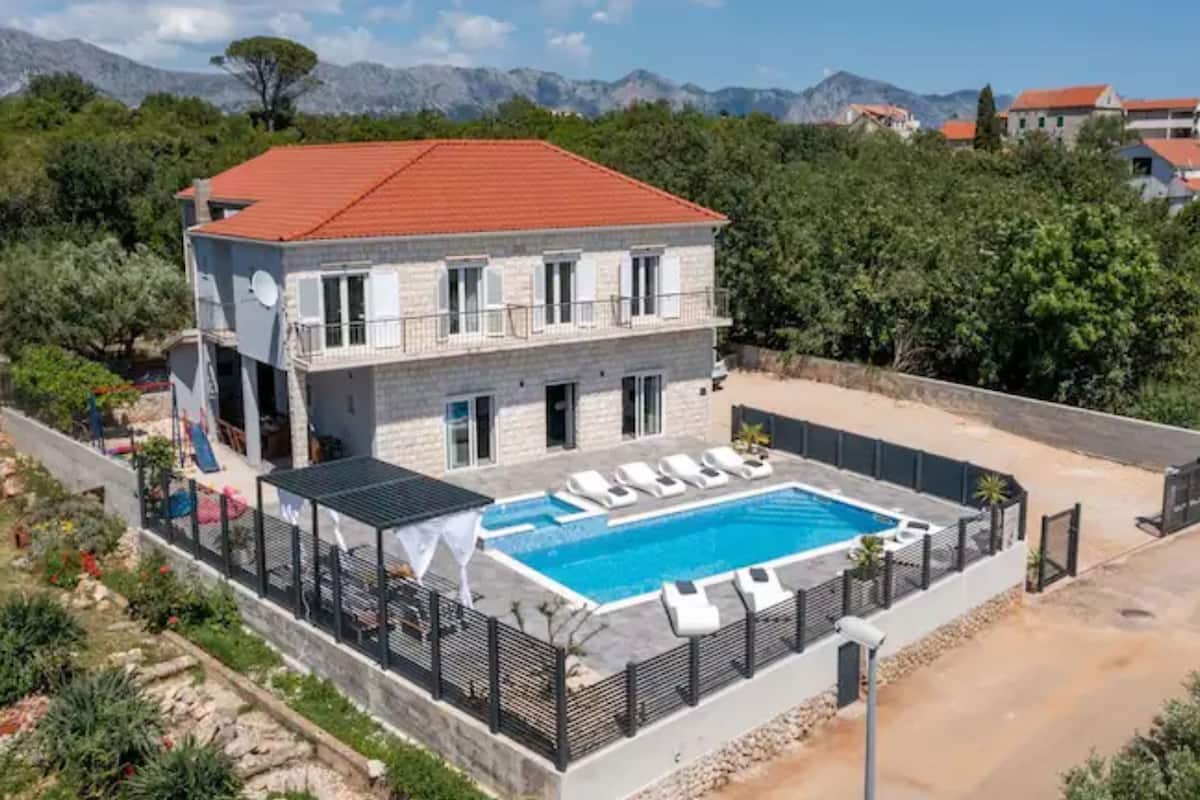 A two-story house with a swimming pool, surrounded by greenery and mountains in the background.