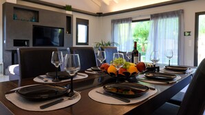 Un coin repas avec une longue table dressée pour quatre personnes, comprenant des verres à vin, des assiettes et des couverts. Un téléviseur est fixé au mur et il y a une fenêtre avec des rideaux.