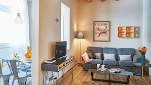 A modern living room with a sofa, coffee table, and a TV. There's a dining area with chairs and a table, and a lamp is on. - Eusebio Planas  - Two Bedroom Apartment, Sleeps 4 (Barcelona)