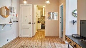 A hallway with a wooden floor, a white door, a kitchen in the background, and a TV on a desk. - Eusebio Planas  - Two Bedroom Apartment, Sleeps 4 (Barcelona)