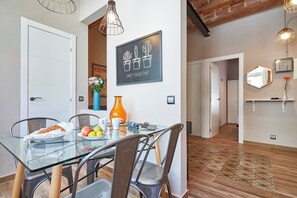 Uma área de refeições com uma mesa de vidro, cadeiras de metal e um prato de comida. Há uma placa emoldurada na parede e um vaso com flores.