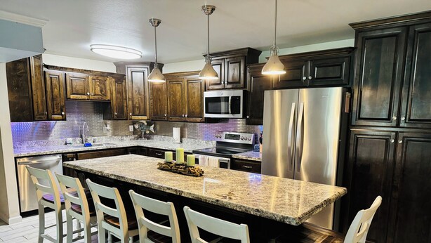 A modern kitchen with dark wood cabinets, a stainless steel refrigerator, a granite countertop island with bar stools, and pendant lighting. - Beautifully Remodeled Condo with 1st Floor Access Steps Away from Beach (South Padre Island)