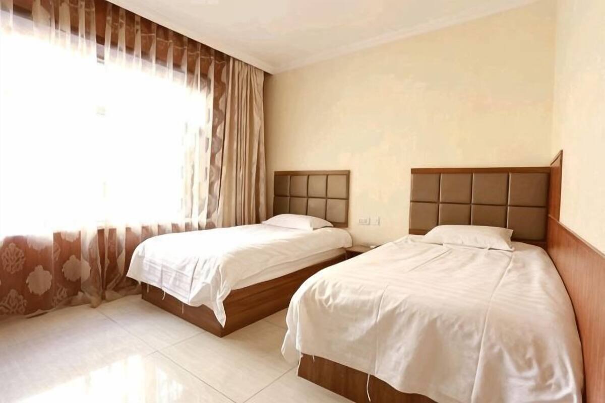 Een hotelkamer met twee bedden, een groot raam met gordijnen en een betegelde vloer.