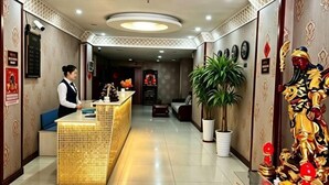 Un espace d’accueil d’hôtel comprenant un comptoir de réception, un réceptionniste, une grande plante en pot et des œuvres d’art décoratives murales.