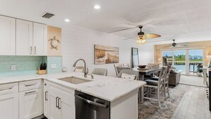 A modern kitchen with white cabinetry, a central island, and a dining area with a view of the outdoors. - Intracoastal-Corner-Pool-Steps to Beach (Indian Shores)
