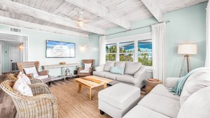 Ein geräumiges Wohnzimmer mit hohen Decken, Korbmöbeln, einem weißen Sofa und Blick ins Freie durch große Fenster.