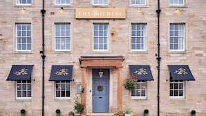 A stone building with a sign reading "THE WILDING" above the entrance. There are three windows with awnings on either side of the door. - Wilding Hotel (Kirkby Stephen)