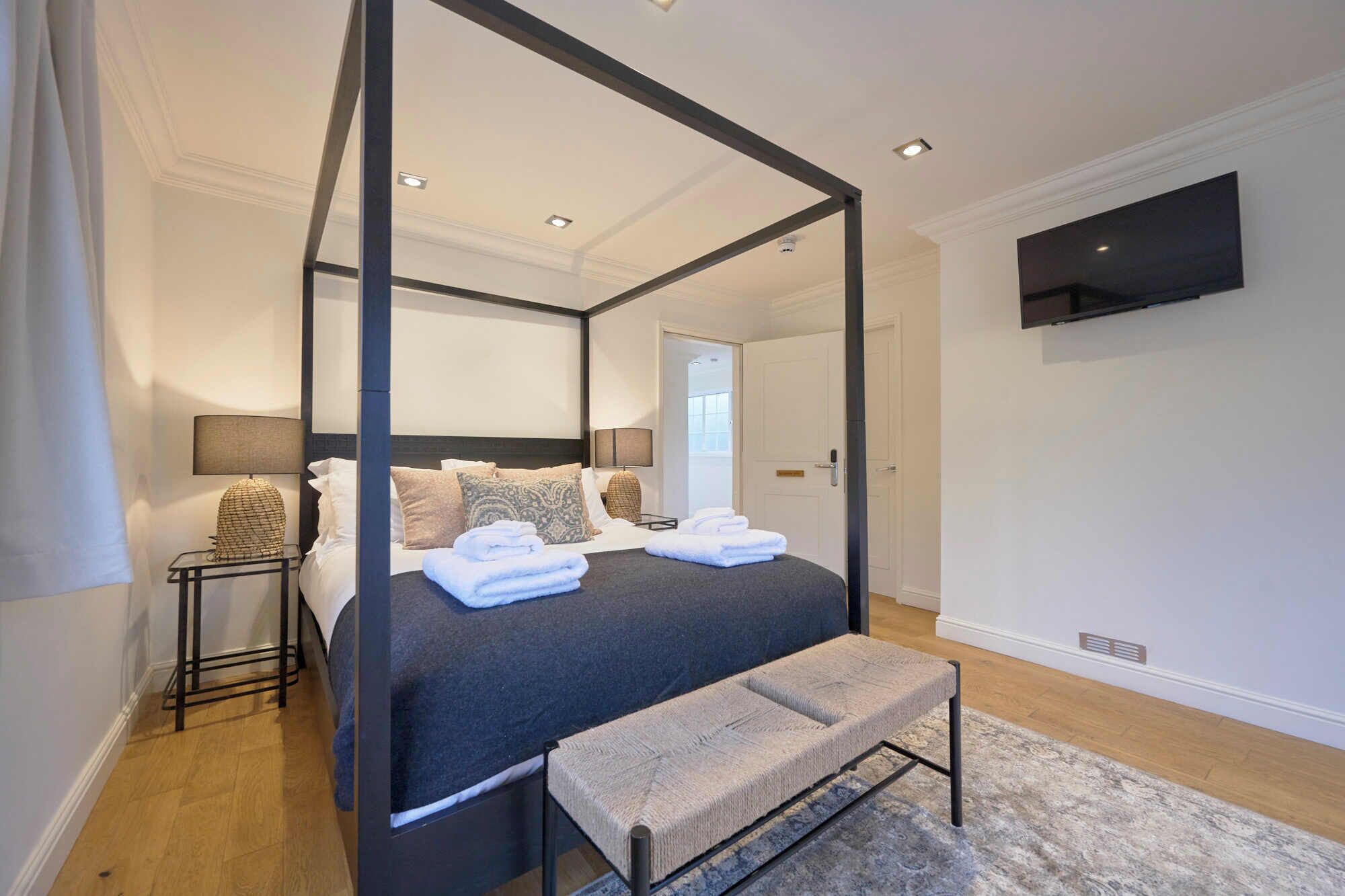 Una cama con dosel, con cabecera oscura, un banco al pie de la cama y un televisor de pantalla plana montado en la pared.