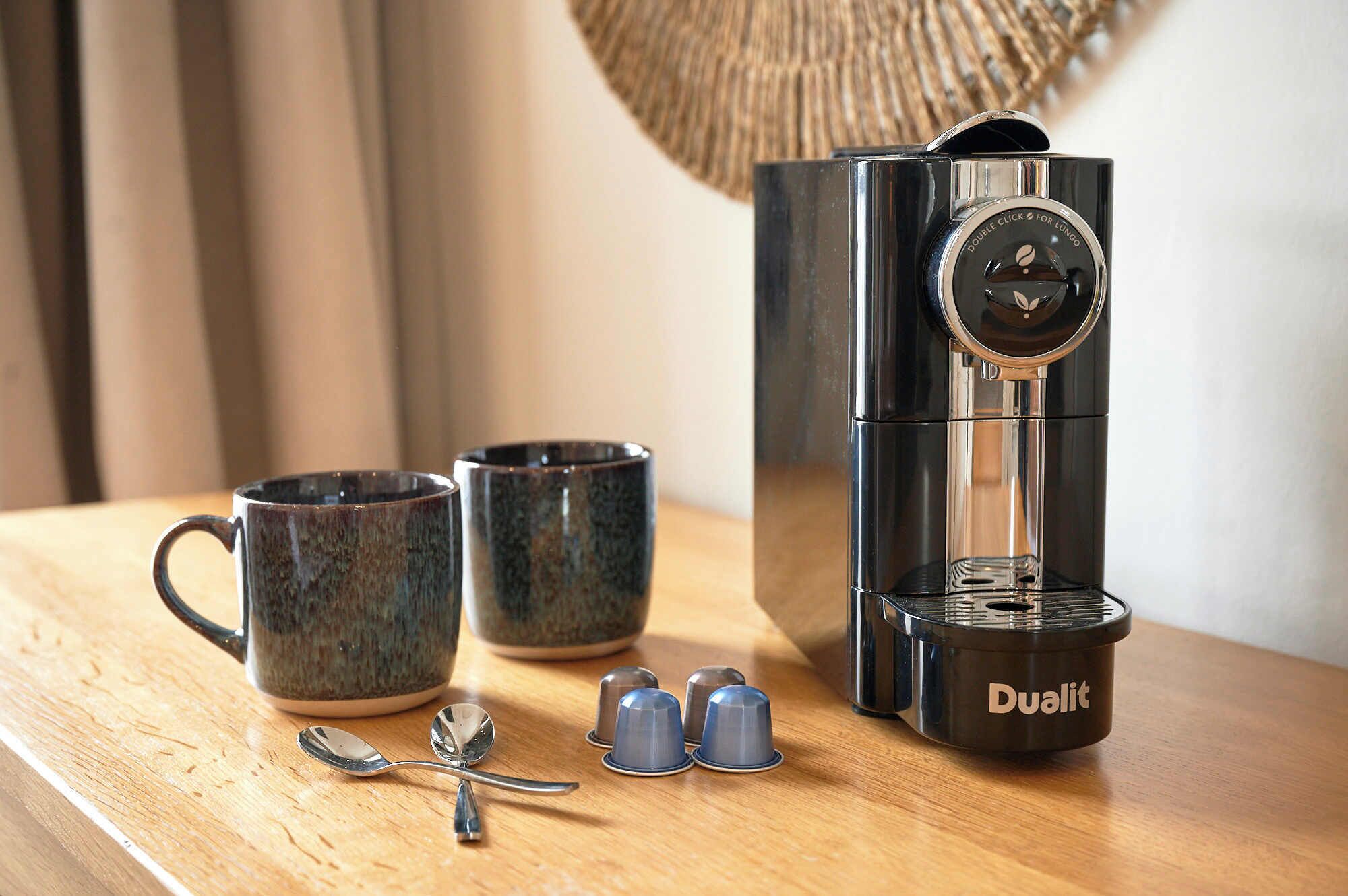 A coffee maker with a Dualit brand, two ceramic mugs, a spoon, and four small blue capsules on a wooden table.
