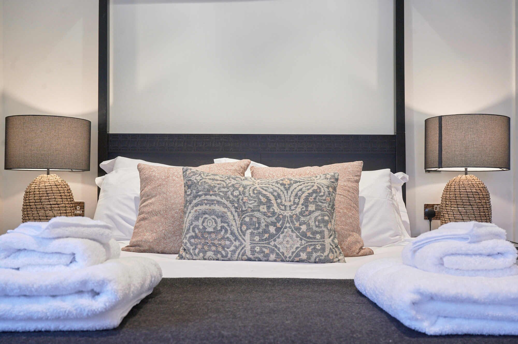 A hotel room with a bed, two bedside lamps, and two rolled towels.