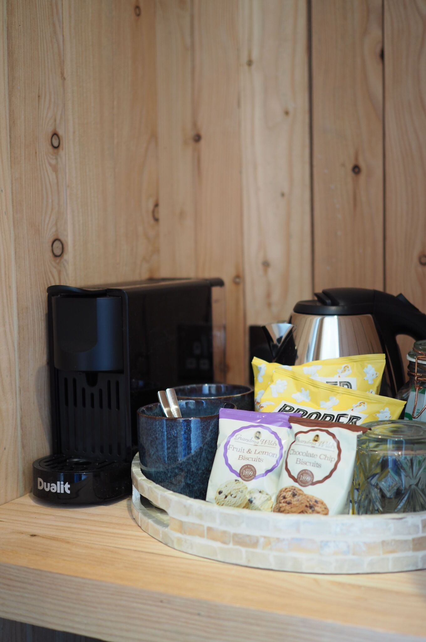A kitchen area with a coffee maker, a kettle, and a tray of food items including biscuits and a bag of coffee.