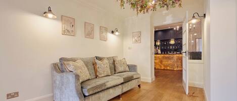 Un salón con sofá gris, suelo de madera, luces fijadas en la pared y un arreglo floral colgante.