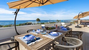 A dining table set with blue and white plates, bowls, and glasses, under a large beige umbrella, overlooking a scenic ocean view.