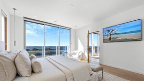 A modern bedroom with a large bed, a view of the sea through floor-to-ceiling windows, and a painting of a tree on the wall.