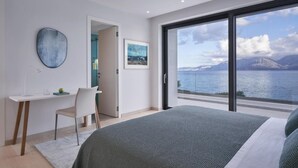 Une chambre avec un grand lit, un bureau et une chaise. Une grande fenêtre offre une vue sur un plan d’eau et des montagnes.