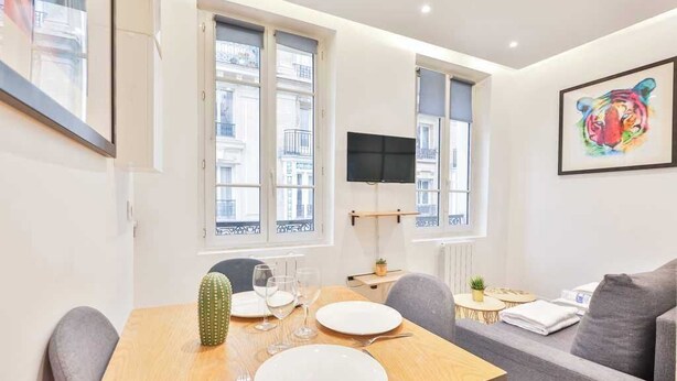 A dining area with a wooden table set for two, a grey sofa, and a TV mounted on the wall.