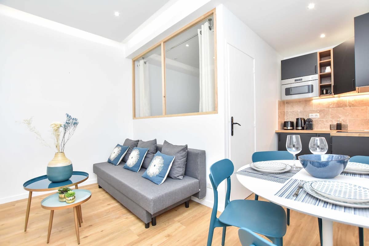A modern living room with a grey sofa, blue chairs, a small round table, and a dining table set for two.