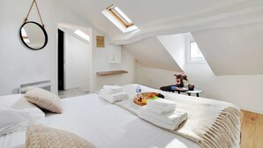 A modern, well-lit bedroom with a skylight, a neatly made bed with towels, a fruit basket, and a small table with a vase.
