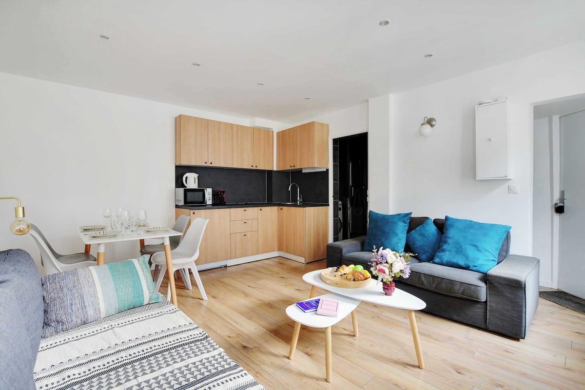 A modern living room with a grey sofa, a dining table with chairs, a kitchen area with wooden cabinets, and a grey couch with blue cushions.