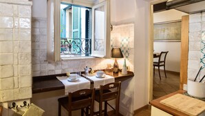 A cozy kitchen with a dining area, featuring a window with open shutters, a table set with cups and a lamp, and a wooden chair. - Estate4home - Trevi Fountain Apartment (Rome)