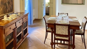 A dining area with a wooden table set for four, flanked by chairs with patterned upholstery. A wooden cabinet with glass doors is to the left, and a painting hangs on the wall above the table. - Estate4home - Trevi Fountain Apartment (Rome)
