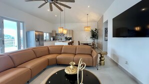 Una sala de estar moderna con un sofá seccional curvo, una mesa redonda de centro con una escultura, un ventilador de techo y una cocina al fondo.